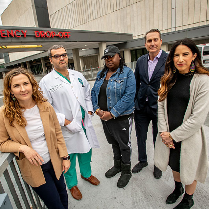 A diverse group of people stand in front of an emergency room drop-off at a hospital.